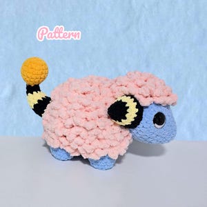 May include: A crochet plush toy shaped like a creature with a light blue face, large black eyes, and a fluffy light pink body. It features a long tail with black and yellow stripes ending in a yellow sphere, and black and yellow ears. The word Pattern is visible.
