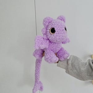 Puede incluir: Un juguete de peluche de dragón lavanda con ojos grandes y redondos y pequeñas alas. El dragón tiene una cola larga y está hecho de un material suave y difuso. El juguete se sostiene sobre un fondo blanco.