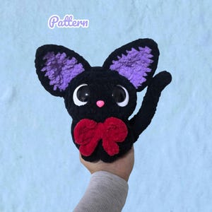 May include: A black crocheted plush toy featuring large purple inner ears, a small pink nose, and two oversized black and white eyes. The creature has a bright red bow tied around its neck and a curved tail. The word Pattern is written above.