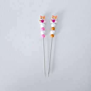 Könnte beinhalten: Zwei dekorative Spieße mit einem Corgi-Hunde-Design, das ein Herz hält. Jeder Spieß hat eine Reihe von weißen, rosa und orangefarbenen Perlen. Die Spieße sind aus Metall und auf weißem Hintergrund.