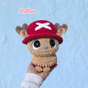 May include: A crochet plush toy of a tan creature with large black eyes, a small blue nose, and brown antlers. It wears a bright red hat featuring a white cross symbol. The word Pattern is written in pink script above the plush toy.