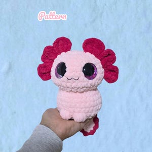 Könnte beinhalten: Ein hellrosa gehäkeltes Axolotl-Stofftier mit dunkelmagentafarbenen äußeren Kiemen und großen, glänzenden schwarzen Augen mit violetten Glitzerakzenten. Das Wort Pattern steht in weißer Schreibschrift über dem Stofftier vor hellblauem Hintergrund.