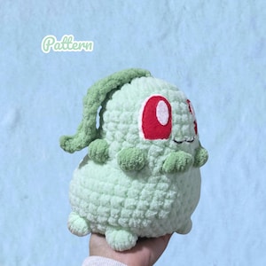 Puede incluir: Un peluche de ganchillo de color verde claro con un cuerpo redondeado, extremidades pequeñas y un largo apéndice curvo en forma de hoja en la parte superior. La criatura tiene grandes ojos rojos ovalados con centros blancos y una pequeña boca cosida. La palabra Pattern es visible.
