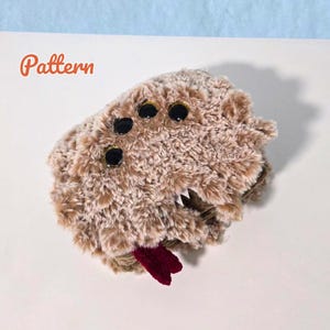 Può includere: Un peluche soffice color marrone chiaro a forma di ragno con molteplici occhi neri, zanne bianche e una lingua rosso scuro. La parola Pattern è stampata in arancione nell'angolo in alto a sinistra dell'immagine, su uno sfondo bianco.