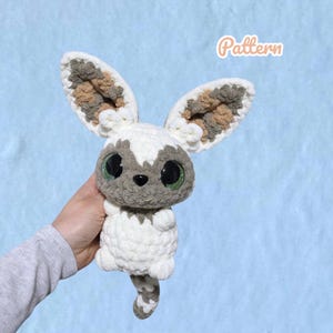 May include: A plush crochet creature with large pointed ears featuring mottled brown, gray, and white yarn. It has a white body, a gray face, large green eyes, and a small black nose. The word Pattern is printed in the upper right corner.