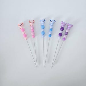 Peut inclure: Cinq épingles décoratives avec des motifs d'axolotl. Chaque épingle présente une petite figurine d'axolotl sur une série de perles colorées roses, bleues et violettes. Les épingles sont disposées sur un fond blanc.