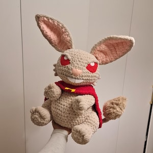 Pu&ograve; includere: Una bambola di coniglio all'uncinetto fatta a mano con grandi orecchie, occhi rossi e un mantello rosso. Il coniglio &egrave; beige con l'interno delle orecchie rosa e una bocca sorridente. Il mantello ha un emblema giallo.