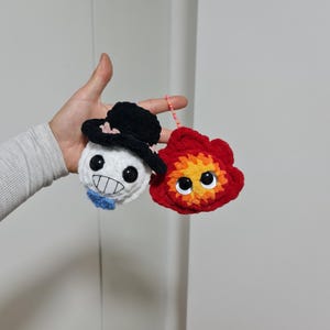 Puede incluir: Dos adornos de personajes de ganchillo. Uno es una calavera blanca con un sombrero de copa negro y una pajarita azul. El otro es una llama roja y naranja con grandes ojos amarillos. Ambos están hechos a mano y conectados por una cuerda rosa.