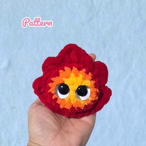 May include: A plush crochet flame character with a deep red outer layer and a bright orange and yellow center. The character features two large, round, black and white plastic eyes. The word "Pattern" is printed in white text in the upper left.