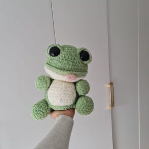 May include: A handmade crochet frog plushie in shades of green and white. The frog has large black eyes, a pink tongue, and a white belly. The plush toy is soft and cuddly, perfect for children or as a decorative item.