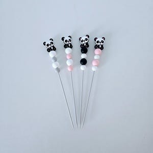 Pode incluir: Quatro espetos decorativos com enfeites de urso panda. Cada espeto apresenta um esquema de cores de contas diferente: cinza, rosa, preto e branco. Os pandas são pretos e brancos com pequenos laços pretos.