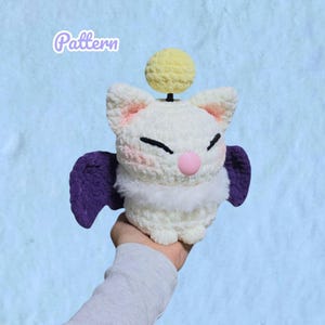 May include: A white crocheted plush toy with purple wings, a pink nose, and a yellow pom-pom on a black antenna. The plush has embroidered black eyes and pink cheeks. The word Pattern is written in white text in the upper left corner of the image.