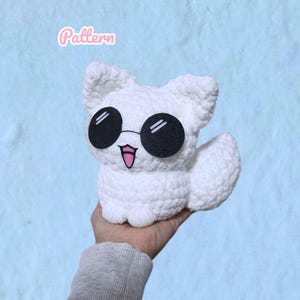 Pode incluir: Um peluche de croché branco em forma de raposa com óculos de sol pretos redondos grandes e uma pequena língua cor-de-rosa de fora. A palavra Pattern está escrita em cursivo cor-de-rosa sobre o peluche, que tem uma superfície de fio texturizada.