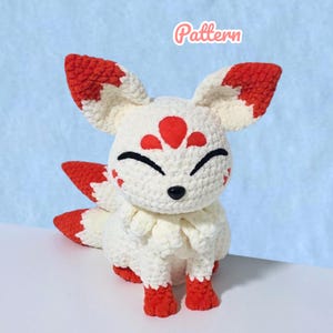 Puede incluir: Un peluche de ganchillo con forma de zorro hecho con hilo blanco y rojo. El zorro tiene orejas grandes y puntiagudas con puntas rojas, una marca roja en la frente, ojos cerrados, nariz negra y varias colas. La palabra Pattern aparece en rosa.
