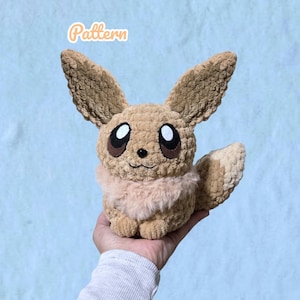 May include: A tan crochet plush toy featuring large pointed ears, a fluffy cream-coloured chest ruff, and dark brown eyes with white highlights. The creature has a small black nose and a stitched mouth. The word Pattern is written in orange text.