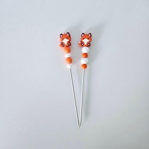 Op de afbeelding: Twee decoratieve spiesen met oranje vossenfiguren. Elke spies heeft oranje en witte kralen. De vossenfiguren zijn oranje met witte en zwarte accenten. De spiesen zijn gemaakt van zilverkleurig metaal.