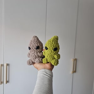 May include: Two small, handmade crochet creatures. One is light brown, the other is green. Both have black eyes and small arms and legs. The creatures are held up against a white background.