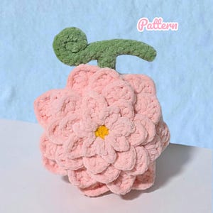 Könnte beinhalten: Eine gehäkelte Plüschblume mit geschichteten hellrosa Blütenblättern, einer kleinen gelben Mitte und einem strukturierten grünen Stiel mit einem eingerollten Blatt an der Spitze. Das Wort Pattern steht in weißer Schreibschrift oben rechts.