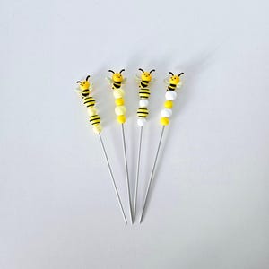 Peut inclure: Quatre brochettes décoratives sur le thème des abeilles. Chaque brochette présente une figurine d'abeille jaune et noire sur une tige perlée avec des perles jaunes, blanches et noires. Les brochettes sont disposées sur un fond blanc.