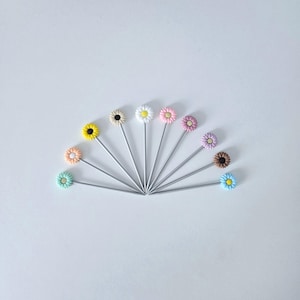 Peut inclure: Une collection de dix épingles décoratives à tête de fleur disposées en éventail. Les têtes de fleurs sont de différentes couleurs, dont le jaune, le noir, le blanc, le rose et le bleu. Les épingles ont des tiges argentées.