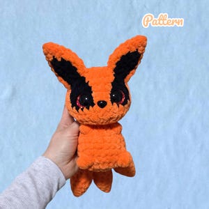 May include: A handmade crochet plush toy featuring a bright orange body with black accents around the large, dark, glittery eyes and pointed ears. The creature has a small black nose and multiple tail-like appendages at the base. The word Pattern is visible.