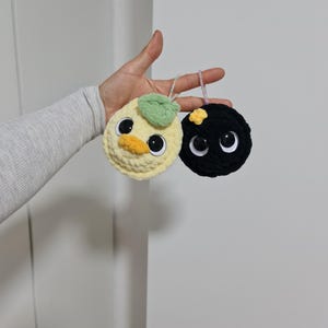 Könnte beinhalten: Zwei gehäkelte Plüschtiere: eine gelbe Ente mit einem grünen Blatt und einem orangefarbenen Schnabel und ein schwarzes Plüschtier mit einer gelben Blume. Beide haben große, runde Kulleraugen und sind an Perlenschnüren aufgehängt.