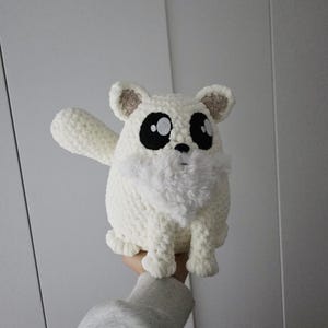 Peut inclure: Un animal en crochet blanc fait à la main, avec de grands yeux noirs et une poitrine blanche duveteuse. Le jouet en peluche a un corps arrondi, de petites oreilles avec des accents beiges et une patte levée. Le jouet est tenu sur un fond blanc.