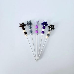 May include: Five bat-shaped cocktail picks. Each pick has a different coloured bat figurine on a beaded stem. The beads are in various colours, including black, grey, purple, and white. The picks are arranged on a white surface.
