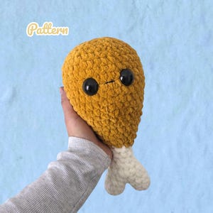 May include: A crochet plush toy shaped like a fried chicken drumstick with a golden-brown textured body, a white bone-shaped base, and two large black plastic safety eyes. The word Pattern is printed in white text in the upper left corner.
