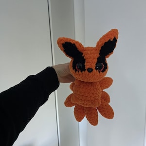 May include: An orange and black crochet plush toy with large ears and multiple tails. The toy has black accents on the ears and around the eyes. The plushie is held against a white background.