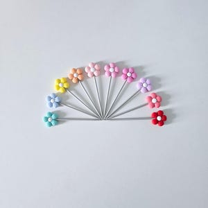 Pode incluir: Um conjunto de palitos de cocktail em forma de flor, dispostos em leque. Cada palito apresenta uma flor de cor pastel diferente, incluindo azul, amarelo, laranja, rosa e vermelho. As hastes são prateadas.
