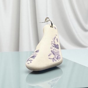 May include: A cream-colored, teardrop-shaped earring with a silver hook. The earring features a purple floral design on its surface. The earring is displayed against a light blue surface and a white backdrop.