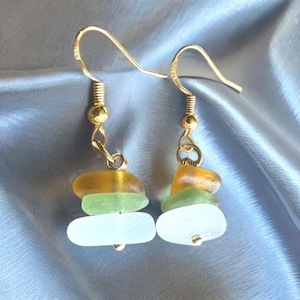May include: Gold-coloured earrings with a layered design. Each earring features three pieces of sea glass in white, green, and amber hues. The earrings have a simple hook closure.