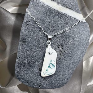 Puede incluir: Un collar de plata con un colgante hecho de un trozo de cerámica blanca con un diseño azul y verde. El colgante está unido a una delicada cadena de plata. El collar se exhibe sobre una piedra gris.