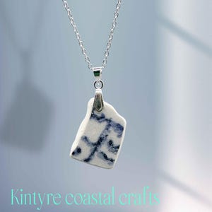 Może przedstawiać: Srebrny naszyjnik z wisiorkiem z białej ceramiki z niebieskimi wzorami. Wisiorek ma prostokątny, lekko nieregularny kształt. Tekst "Kintyre coastal crafts" jest widoczny na dole obrazu.