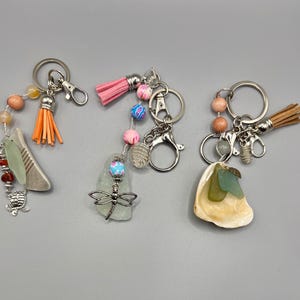 May include: Three handcrafted keyrings with distinctive designs. One has orange tassels and a stone pendant, another a pink tassel and dragonfly charm, and the third a seashell and tan tassel. Each keyring has a silver ring.