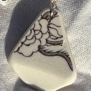 May include: A white, triangular pendant with a black floral design. The pendant is suspended from a silver wire loop. The design is a stylized floral pattern. The pendant is likely made of ceramic or porcelain.