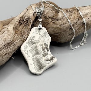 Könnte beinhalten: Eine silberne Halskette mit einem Anhänger aus weißer Keramik. Der Anhänger hat ein schwarz-graues Blumendesign. Die Halskette hat eine zarte silberne Kette.