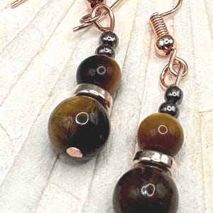 May include: Handmade earrings with tiger's eye and hematite beads. The earrings have copper-coloured hooks and connecting elements. The tiger's eye beads are brown with natural patterns, and the hematite beads are dark grey. A stylish accessory.
