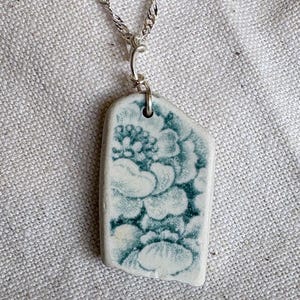 May include: A silver chain necklace with a pendant. The pendant is a rectangular piece of white ceramic with a teal floral pattern. The chain is a twisted rope design.