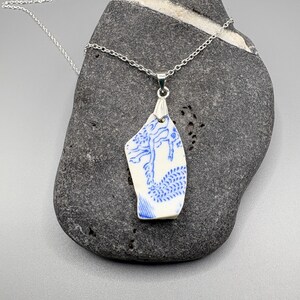 Pode incluir: Um colar de prata com um pingente feito de um pedaço de porcelana branca com um desenho de dragão azul. O pingente, de forma irregular, pende de uma delicada corrente de prata. O fragmento de porcelana é exibido sobre uma pedra cinza escura.