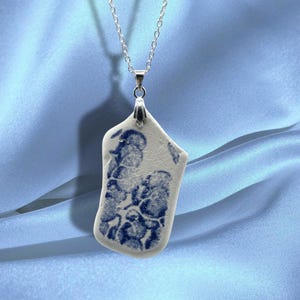 Pode incluir: Colar de corrente prateada com um pendente de cerâmica branca. A cerâmica apresenta um padrão floral azul. O pendente, de forma irregular, destaca-se num fundo azul claro.