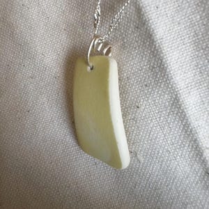 Può includere: Collana con un ciondolo rettangolare verde pallido con un bordo curvo. Il ciondolo è appeso a una delicata catena d'argento. Lo sfondo è un tessuto chiaro e strutturato.