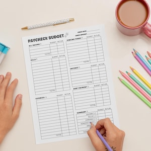 May include: A white paycheck budget sheet with the title "PAYCHECK BUDGET" and sections for income, bills, expenses, and savings. A pen and colored pencils are on the table. A hand is holding a purple pencil.