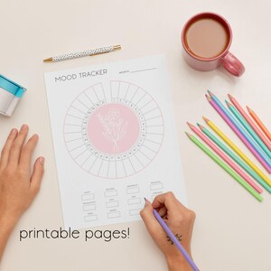 K&ouml;nnte beinhalten: Ein wei&szlig;es Stimmungstracker-Blatt mit einem rosa Blumendesign, einem Stift, Buntstiften und einer Tasse Kaffee. Die W&ouml;rter "Mood Tracker" und "printable pages!" sind sichtbar. Eine Hand schreibt mit einem lila Bleistift auf das Blatt.