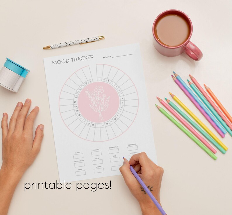 K&ouml;nnte beinhalten: Ein wei&szlig;es Stimmungstracker-Blatt mit einem rosa Blumendesign, einem Stift, Buntstiften und einer Tasse Kaffee. Die W&ouml;rter "Mood Tracker" und "printable pages!" sind sichtbar. Eine Hand schreibt mit einem lila Bleistift auf das Blatt.
