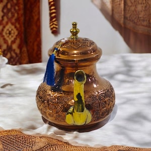 Peut inclure: Une théière en cuivre avec des motifs gravés complexes et un bec verseur couleur or. La théière a un pompon bleu attaché au couvercle.