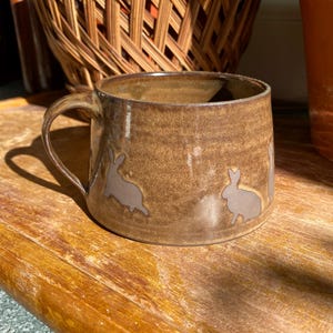 Puede incluir: Una taza de cerámica marrón con asa, con un diseño de siluetas de conejos de color marrón claro. La taza está sobre una superficie de madera, con una cesta de mimbre y una maceta de terracota en el fondo. La taza tiene unos 10 cm de diámetro.