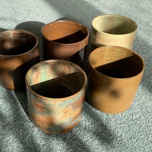 Puede incluir: Cinco tazas de cerámica hechas a mano en tonos marrones y verdes. Las tazas tienen forma cilíndrica con una superficie ligeramente texturizada. Los colores varían desde un marrón oscuro intenso hasta un tono terroso más claro, con toques verdes y azules.
