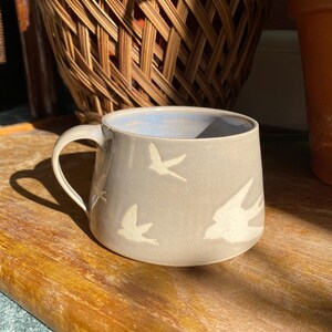 Puede incluir: Una taza de cerámica gris con interior azul claro y asa curva. La taza presenta siluetas de pájaros blancos. La taza está sobre una superficie de madera.